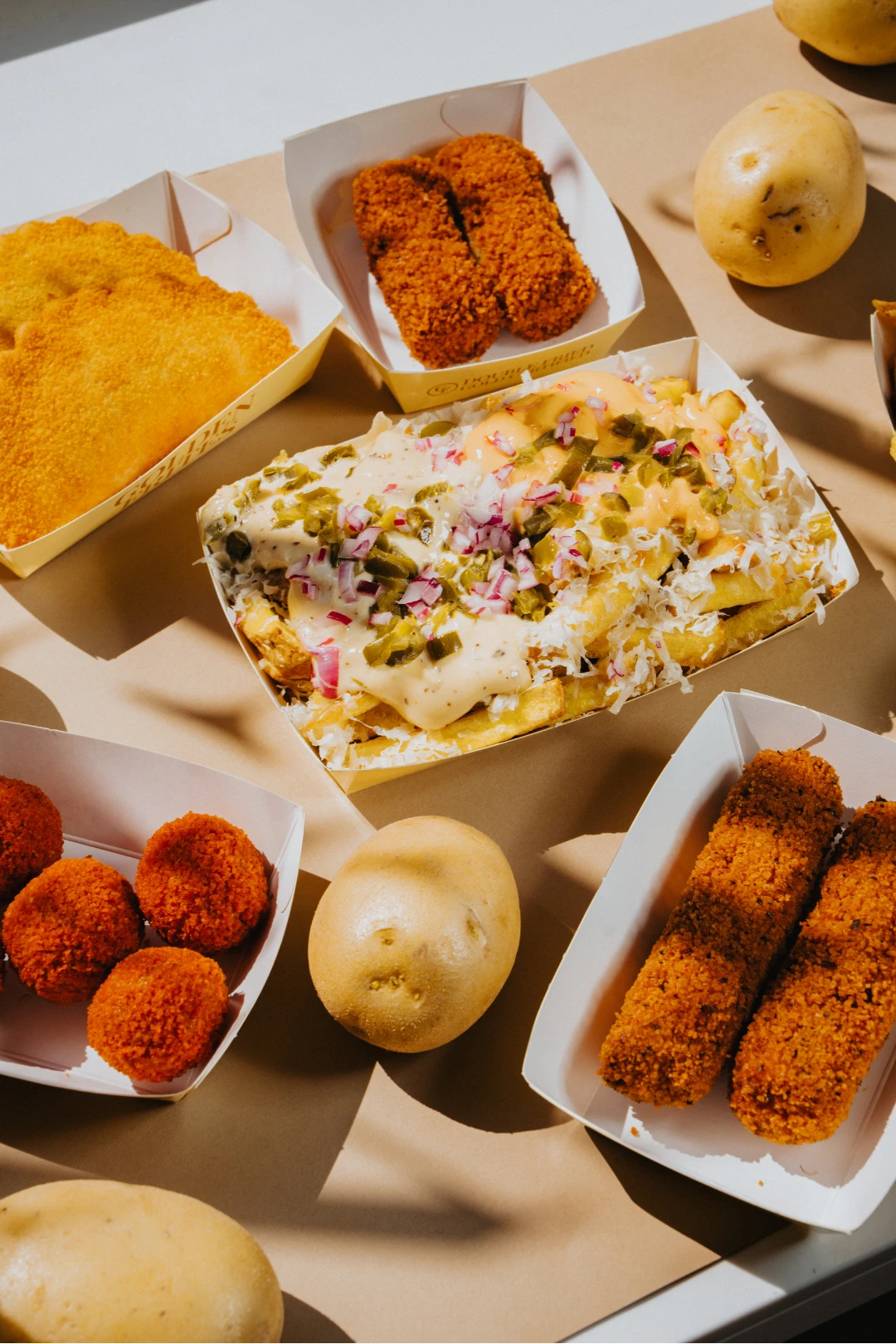 Verschiedene Fast-Food-Gerichte wie Pommes Frites mit Soße und Jalapeños, Kroketten und frische Kartoffeln auf braunem Papier.